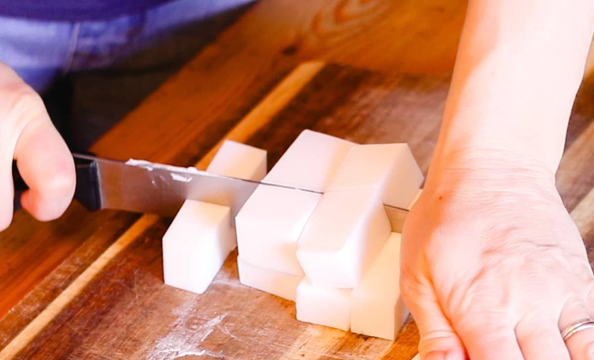 Two hands using a large knife to cut melt-and-pour soap base into roughly equal-sized cubes.
