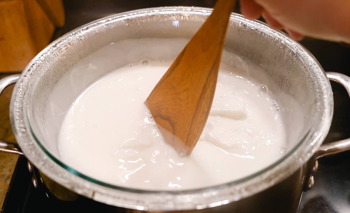 A glass bowl is filled with white melting soap base, set over another large pot of boiling water to make a double boiler. A wooden spoon is stirring the mixture as it melts.