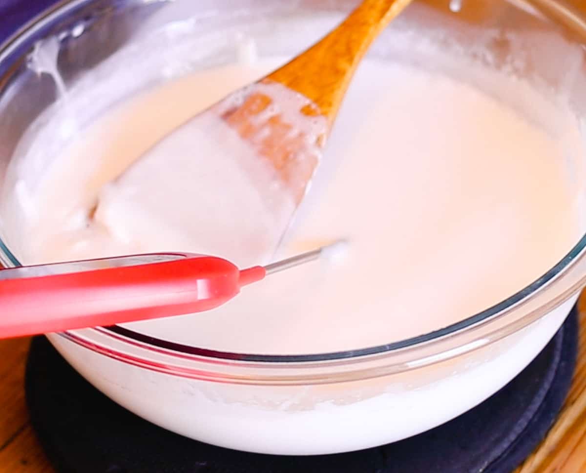 A red instant thermometer dipped into a large glass bowl filled with melted soap base with a wooden spoon mixing the base as it melts.