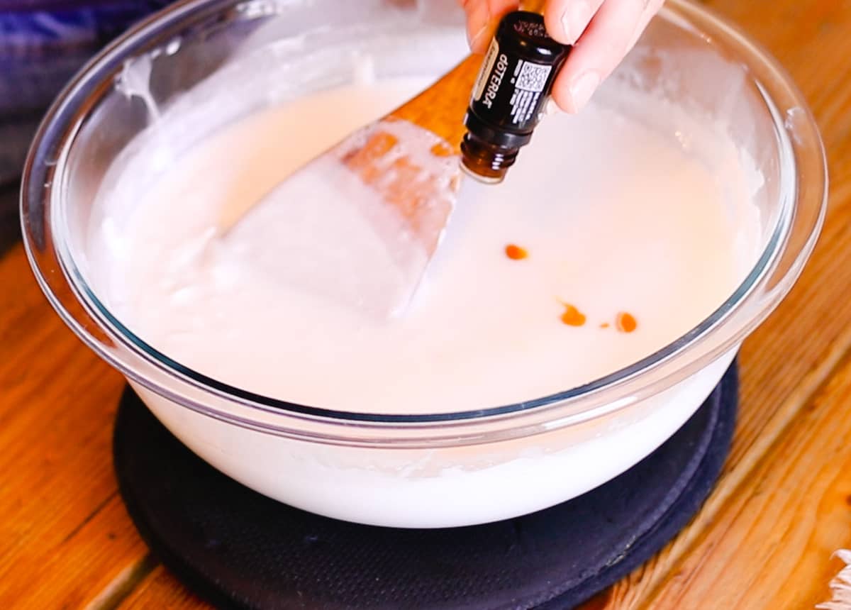 A hand pouring drops of doTERRA essential oil into a large glass bowl filled with melted soap base.