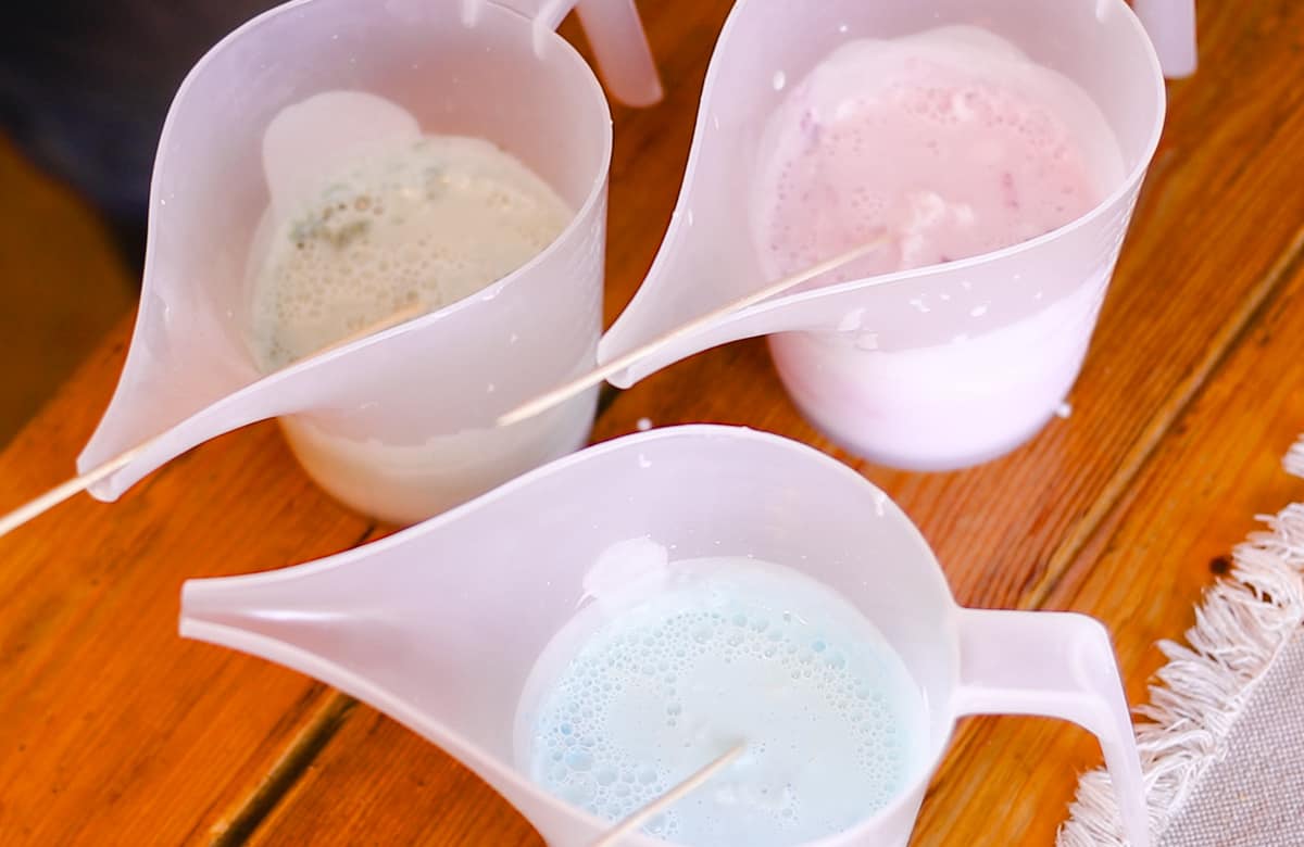 Three plastic pitchers, one green, one pink, and one blue, full of the liquid mixture for homemade bar soap. Each pitcher has a wooden stirring skewer inside the liquid resting against the inner edge of the pitcher.