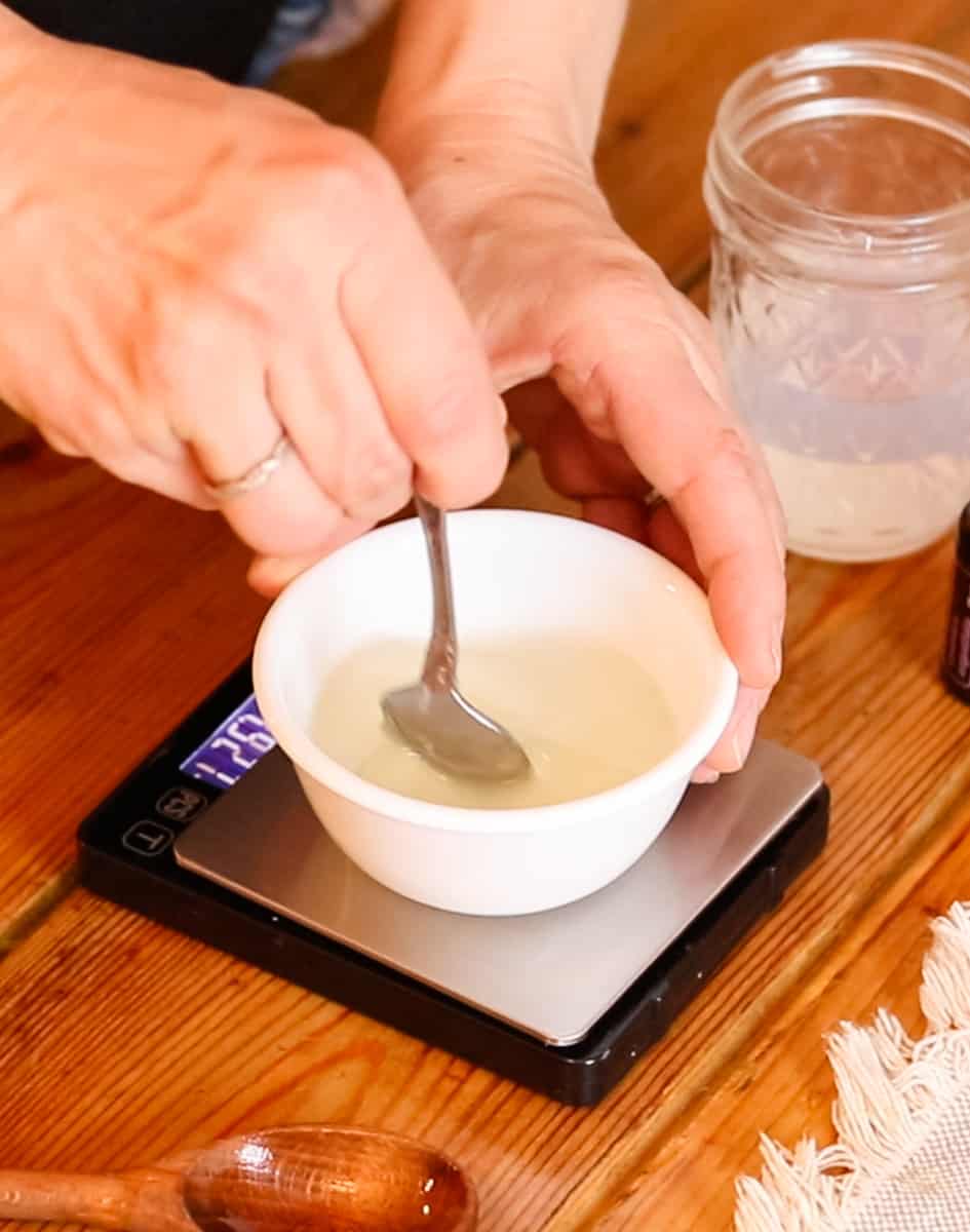 Using a metal spoon to stir a mixture of vegetable glycerin, castile soap, and lavender essential oil in a small bowl on top of a digital scale.