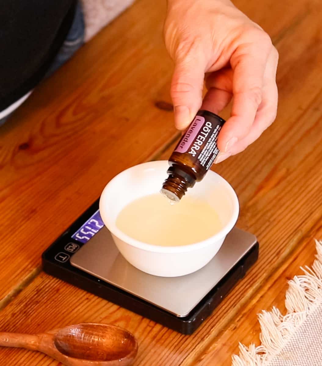 Pouring drops of lavender essential oil into a small bowl containing a mixture of vegetable glycerin and castile soap. The bowl is sitting on a digital scale.