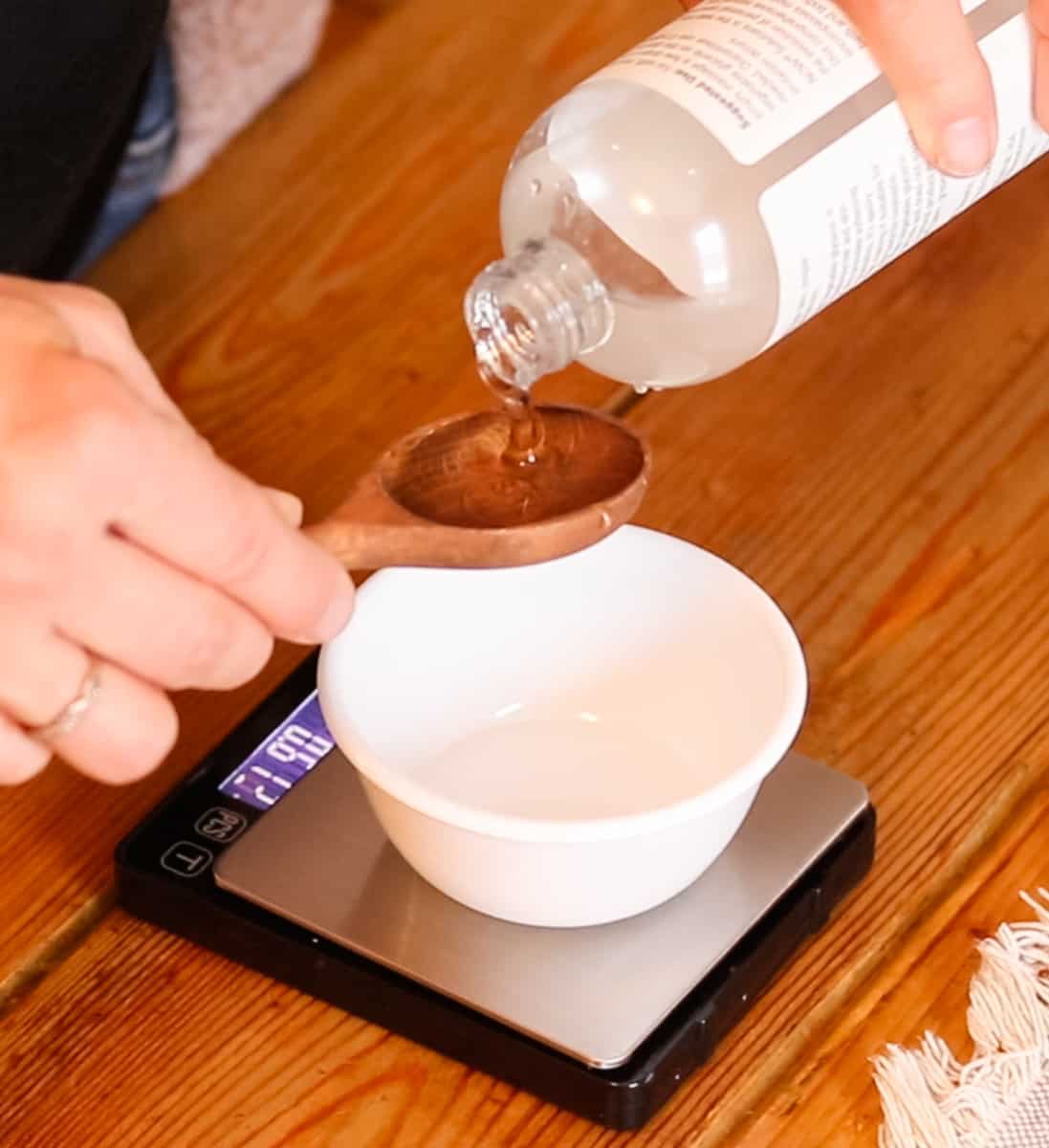 Pouring vegetable glycerin into a wooden spoon held over a small bowl that is sat on top of a digital scale.