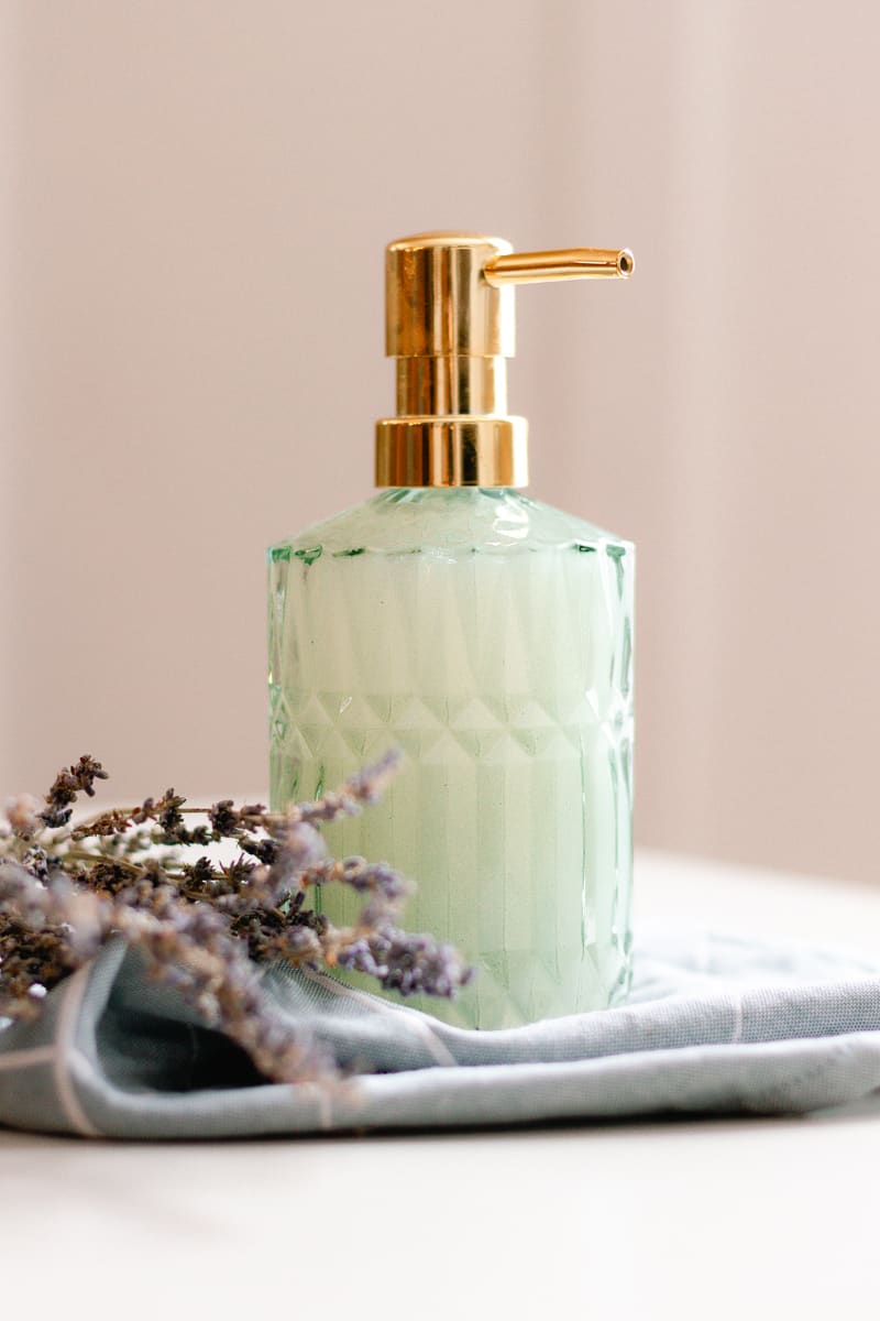 Homemade liquid hand soap in a green tinted embossed glass soap dispenser on top of a folded towel with dried flowers lying beside it.