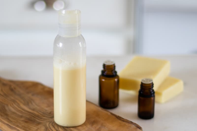 A bottle of coconut rosemary shampoo on a wooden shelf with shampoo bars and 2 essential oil bottle behind it.