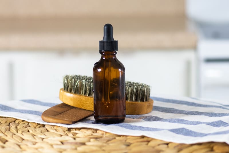 Homemade beard shampoo in an ambered dropper bottle with a beard brush and comb in the background.