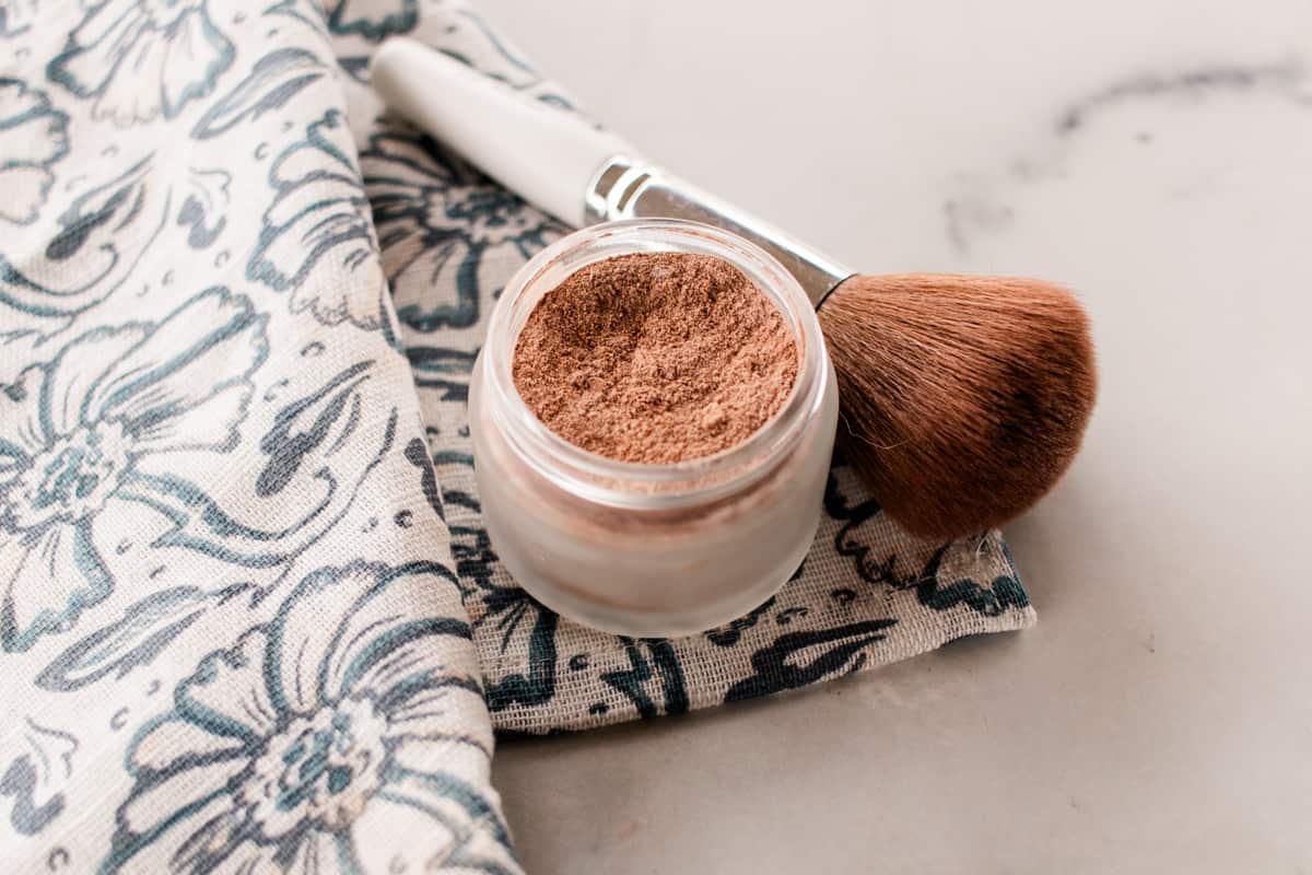 A small glass jar filled with DIY makeup powder with a makeup brush set beside it on a floral piece of material.