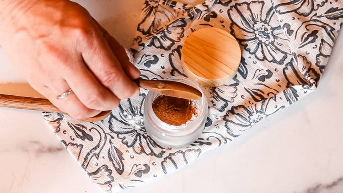 A woman using a wooden spoon to add cocoa powder, cinnamon, and arrowroot powder to a small glass container.