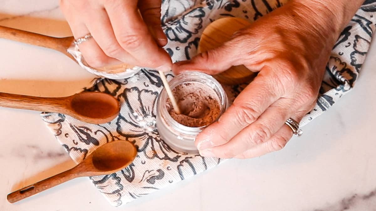 A woman using a wooden stick to mix together ingredients for DIY bronzer in a small glass container.