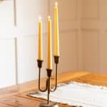 Three beeswax taper candles burning in a bronze candle holder on a wooden table.