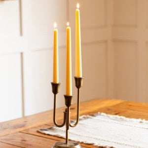 Three beeswax taper candles burning in a bronze candle holder on a wooden table.