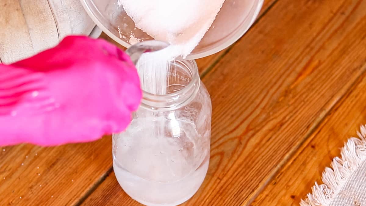 A gloved hand spooning lye into a mason jar filled with water.