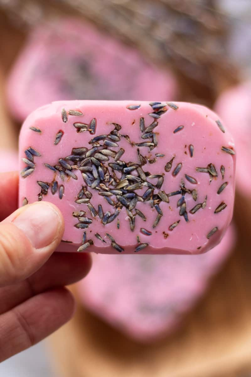 A hand holding a bar of pink soap that has lavender buds pressed into it.