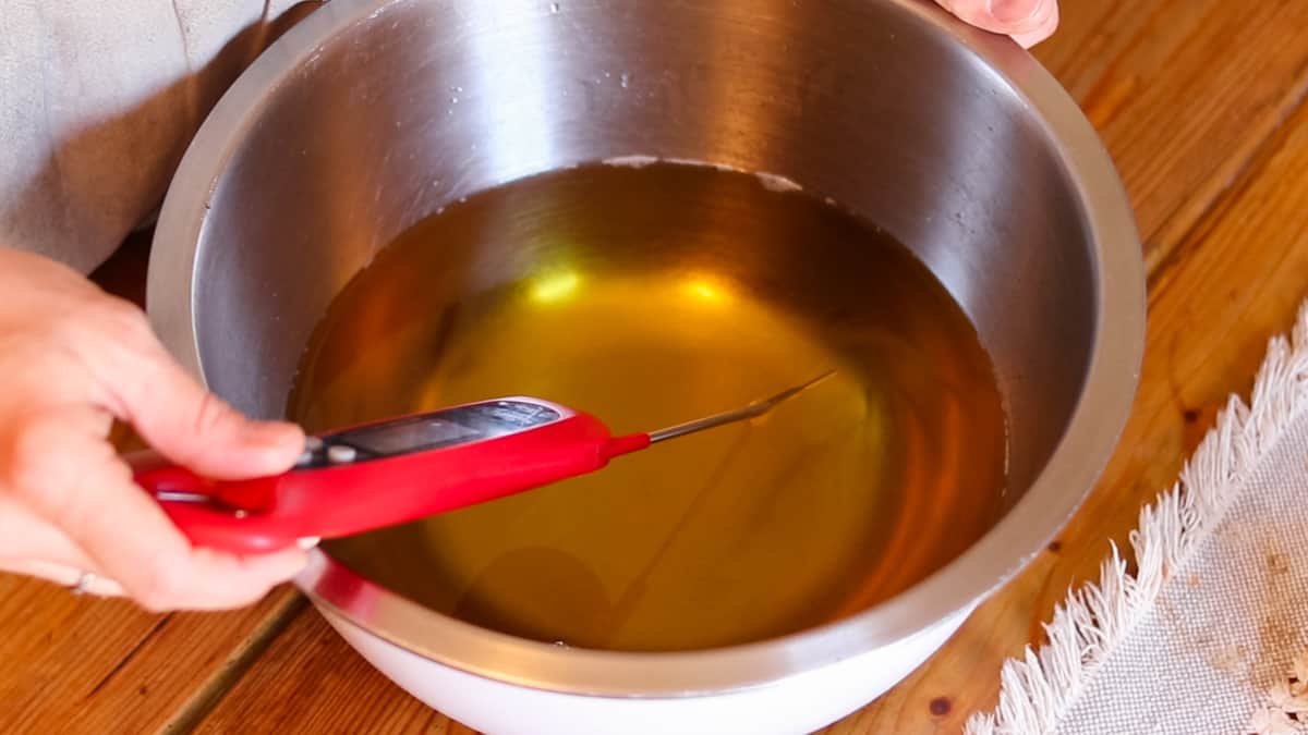 A hand holding a digital thermometer dipped in a bowl of melted oils checking the temperature of the oils.