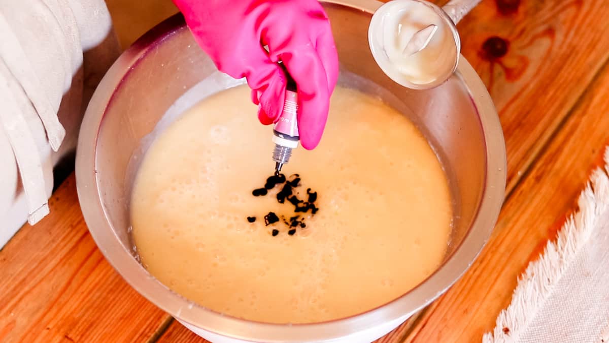 A hand pouring drops of coloring dye into a bowl filled with DIY soap batter.