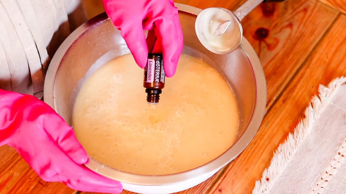A woman pouring lavender essential oil drops into a large bowl filled with soap batter.