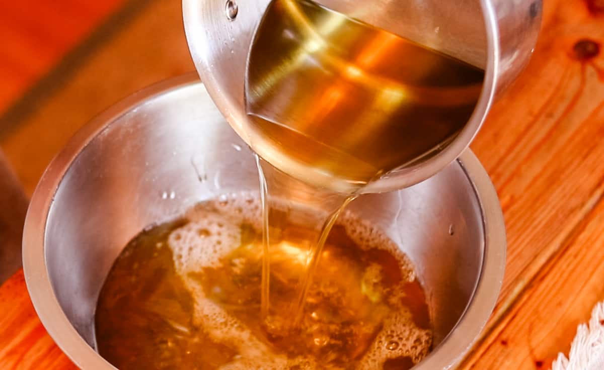 A saucepan filled with melted oils for soap making being poured into a large metal bowl.