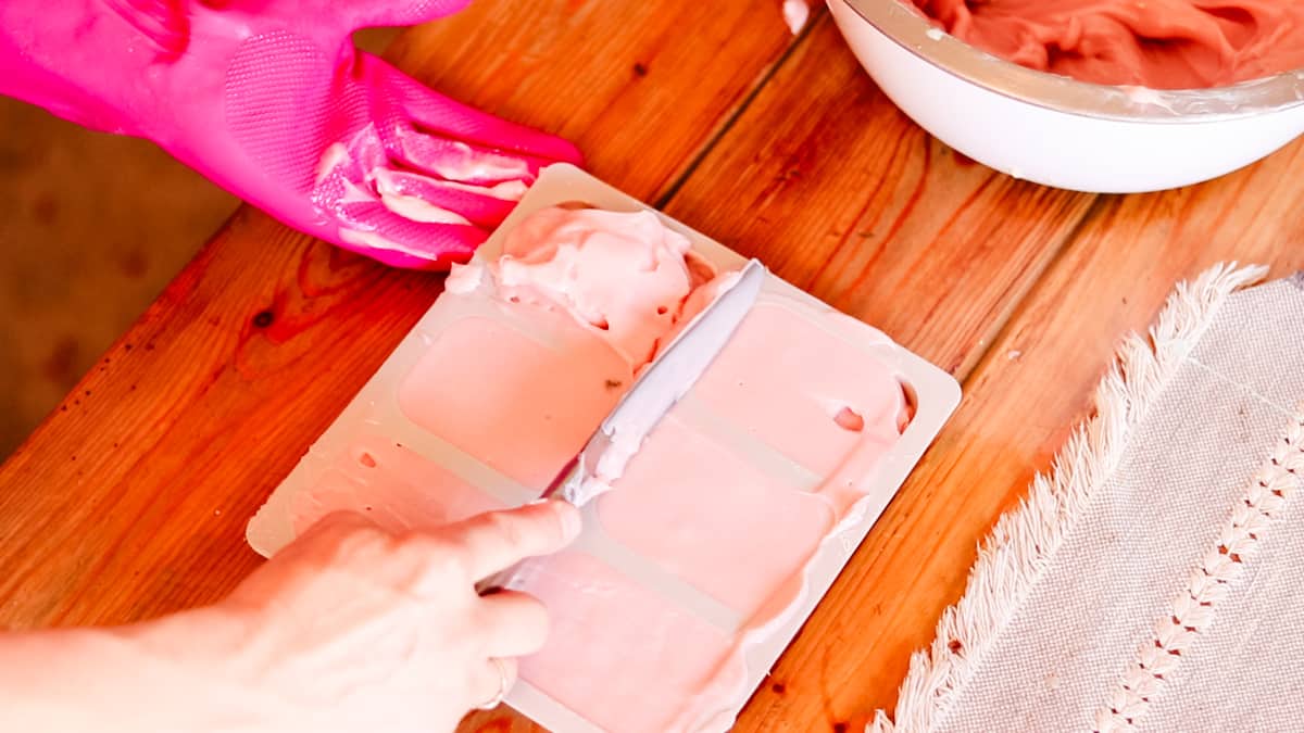 A woman using a metal butter knife to scrape excess soap batter from the top of a silicone soap mold.