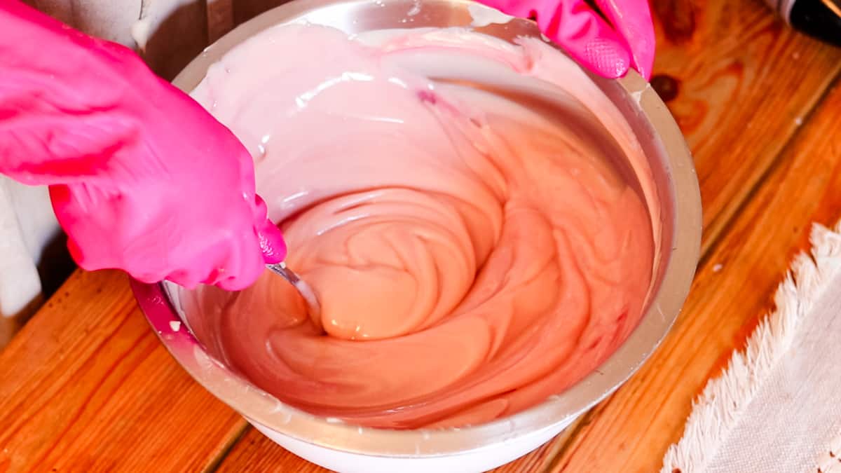 A woman wearing rubber gloves using a metal spoon to stir pink homemade soap batter in a large metal bow.