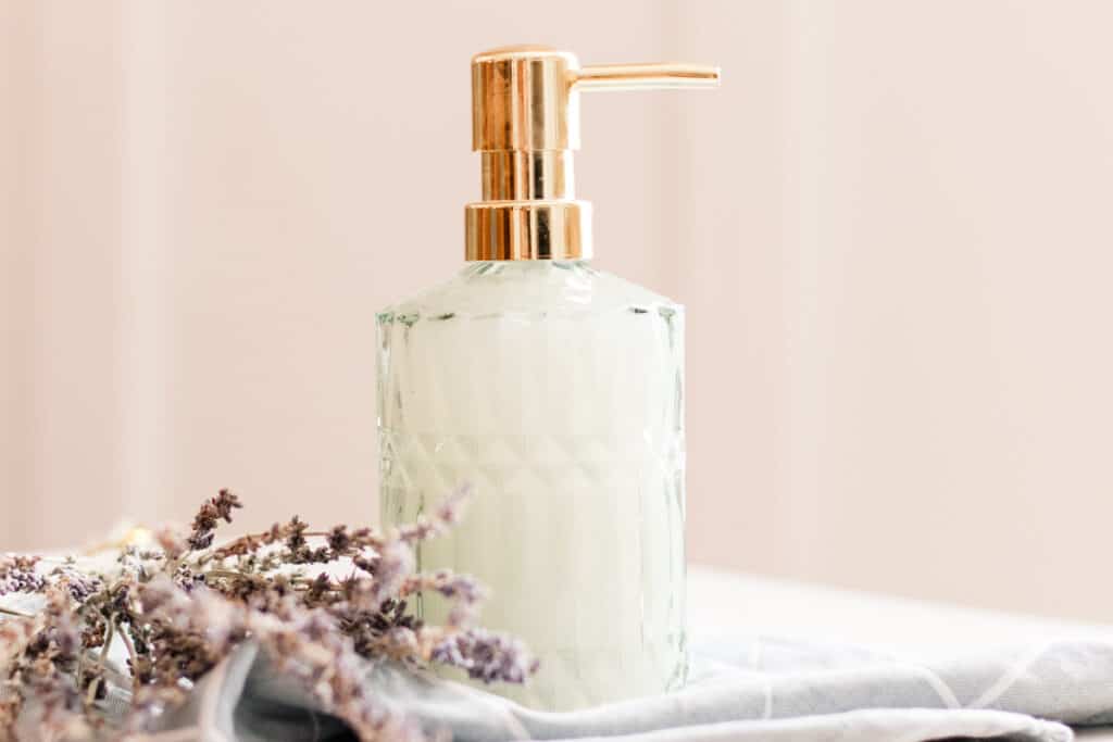 Homemade liquid hand soap in a green tinted embossed glass soap dispenser on top of a folded towel with dried flowers lying beside it.