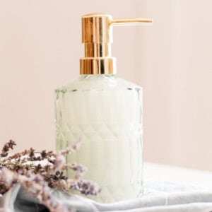 Homemade liquid hand soap in a green tinted embossed glass soap dispenser on top of a folded towel with dried flowers lying beside it.