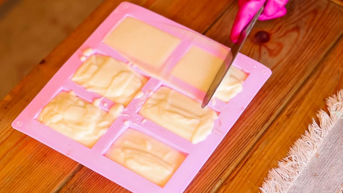 A hand using a metal butter knife to scrape excess batter and level off the tops of soap bars in a silicone mold.