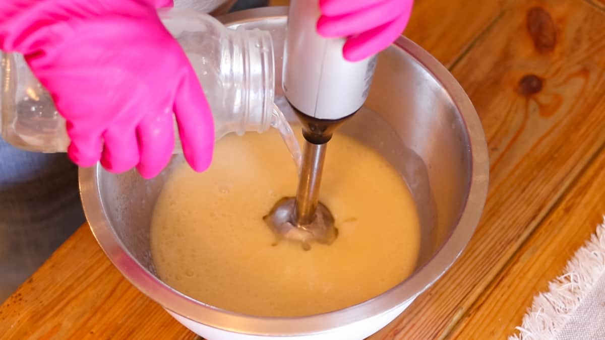 A gloved woman mixing soap ingredients in a large bowl with an immersion blender while pouring lye solution into the mix.