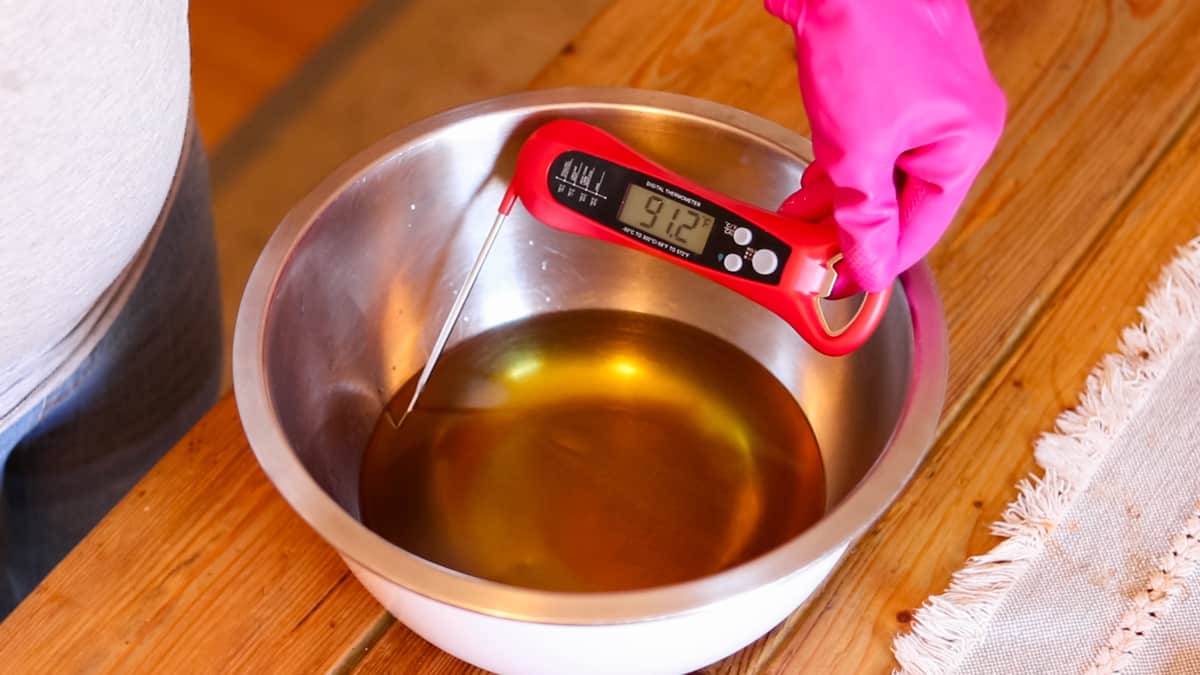 A gloved hand checking the temperature of melted oils in a metal bowl using a digital thermometer.