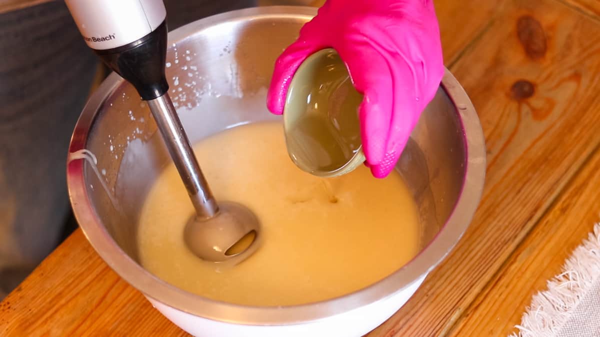 A woman adding fragrance into a bowl with soap ingredients at a light trace with an immersion blender in her other hand.
