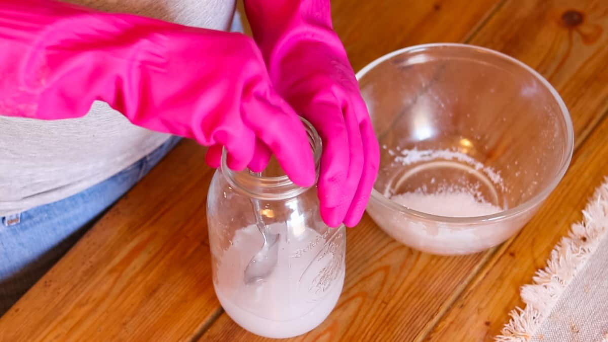 A woman wearing pink rubber gloves stirring a mixture of lye and water with a metal spoon.