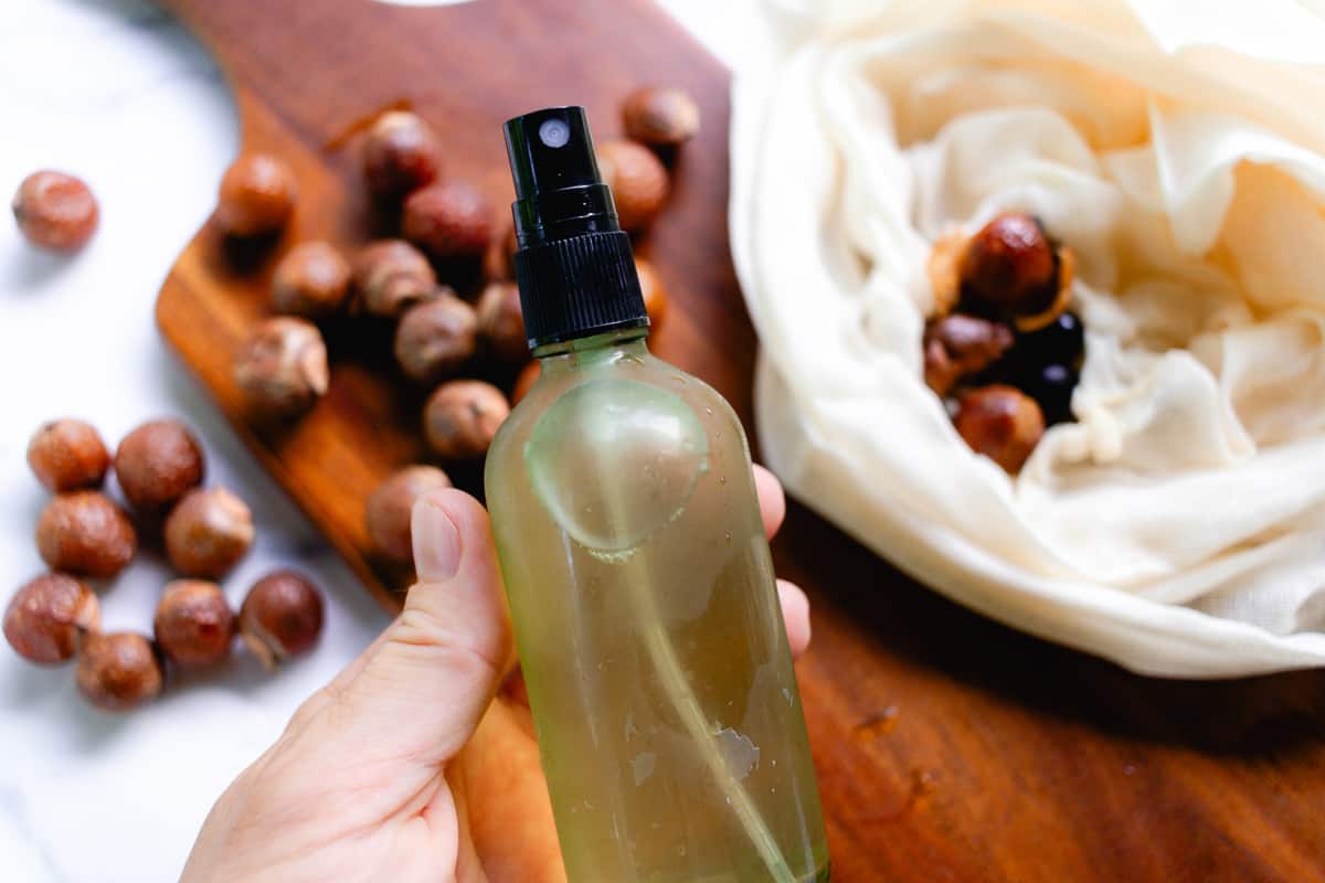 Holding up a small bottle of soap nut shampoo while fresh soap nuts lay on the table below it.