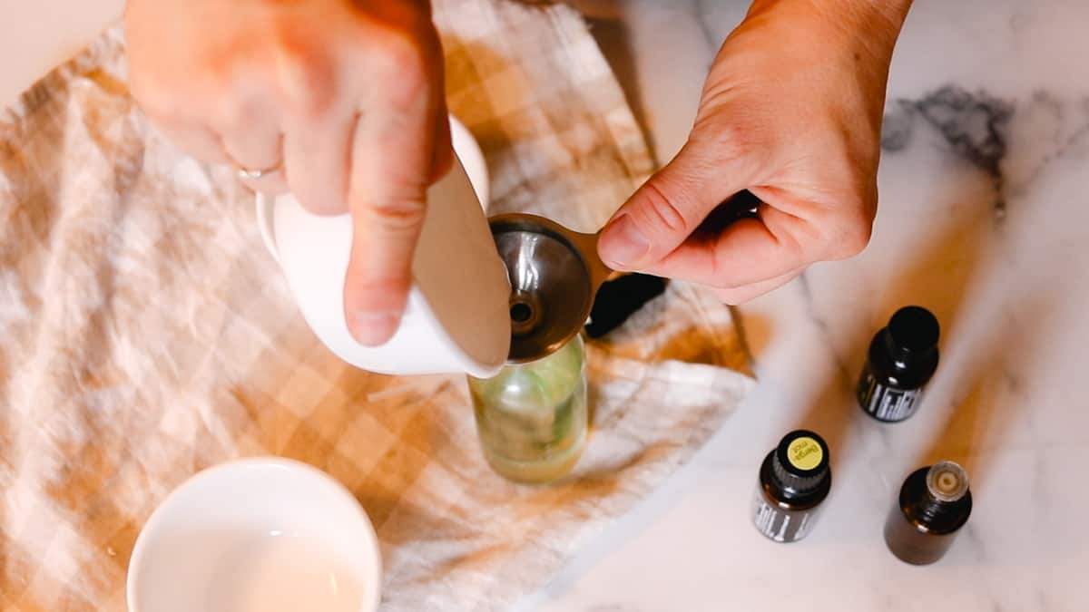 A woman making deodorant by pouring coconut oil through a funnel into a small glass spray bottle.