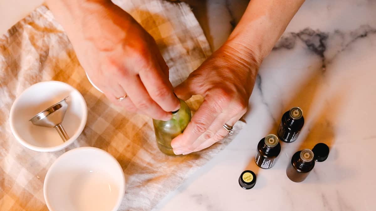 A woman securing the lid of a deodorant spray bottle with essential oils ingredients set nearby on a countertop.