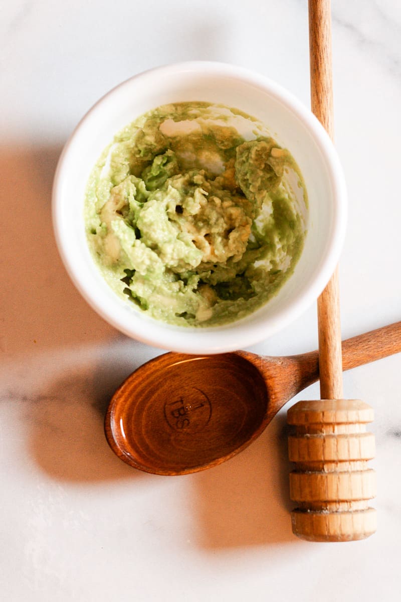 Mashed avocado in a small white ceramic bowl with one tablespoon of honey and a wooden honey stirrer on a white and gray countertop. These are ingredients for a DIY Hair Mask for Curly Hair.