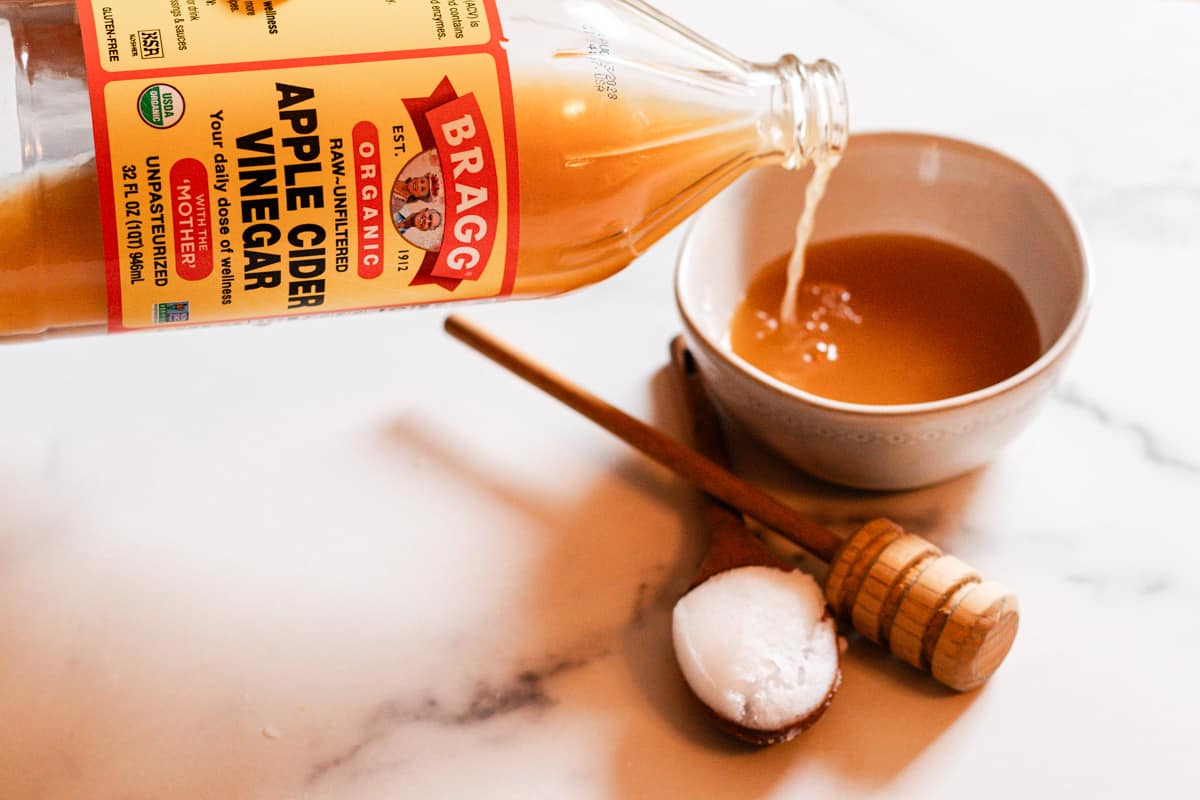 Braggs Apple Cider Vinegar being poured into a small whiite bowl on a white and gray counterop. There is a wooden spoon with coconut oil and a wooden honey stirrer to complete the ingredients for a DIY Hair Mask for Curly Hair. 