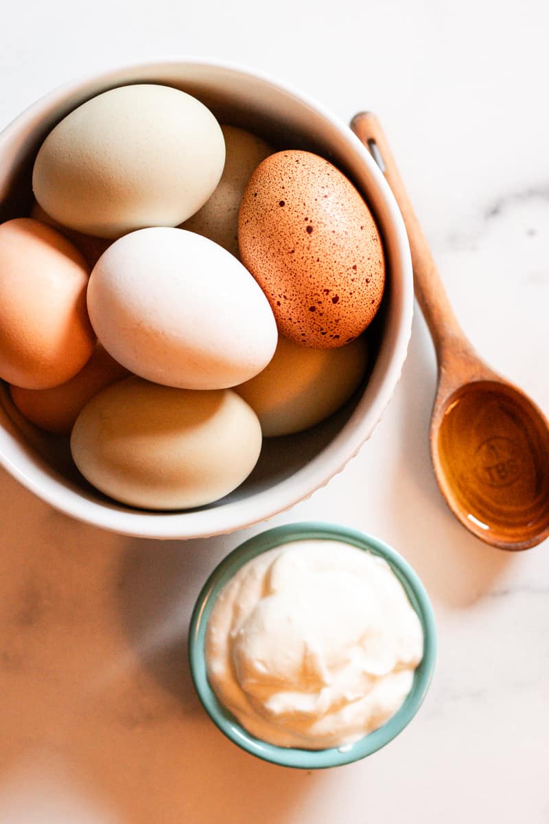A white bowl with several, multicolored and speckled eggs sits on a white and gray countertop next to a wooden spoon filled with honey to make a DIY mask for curly hair.