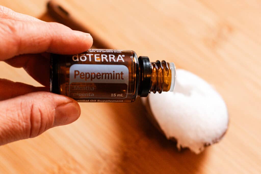 A hand holding a bottle of DoTerra peppermint oil over a wooden spoon of coconut oil. These ingredients are used for a DIY hair mask for curly hair.