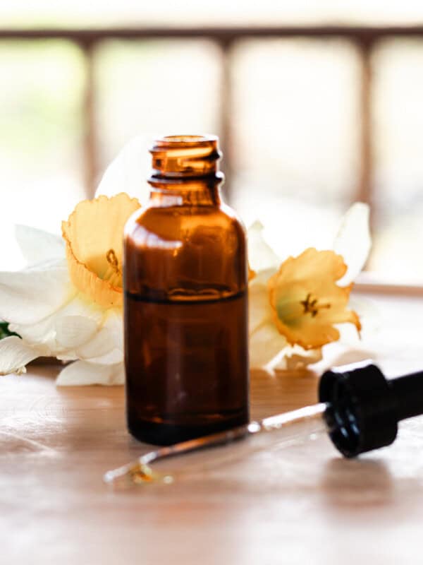 A brown glass bottle filled with homemade dry scalp serum. The dropper lid lies on the table in front and there is a daffodil in the background for decoration.
