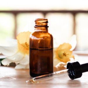 A brown glass bottle filled with homemade dry scalp serum. The dropper lid lies on the table in front and there is a daffodil in the background for decoration.