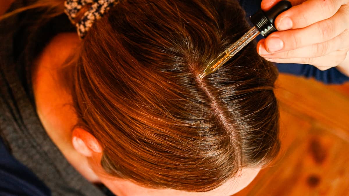 A woman with brown hair uses a dropper to apply a homemade dry scalp hair serum to her scalp. 