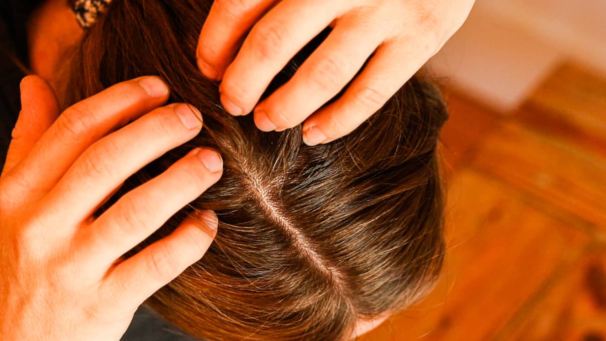Two hands pull apart brown hair to expose the scalp on a woman's head before treating the scalp with a homemade dry scalp serum.