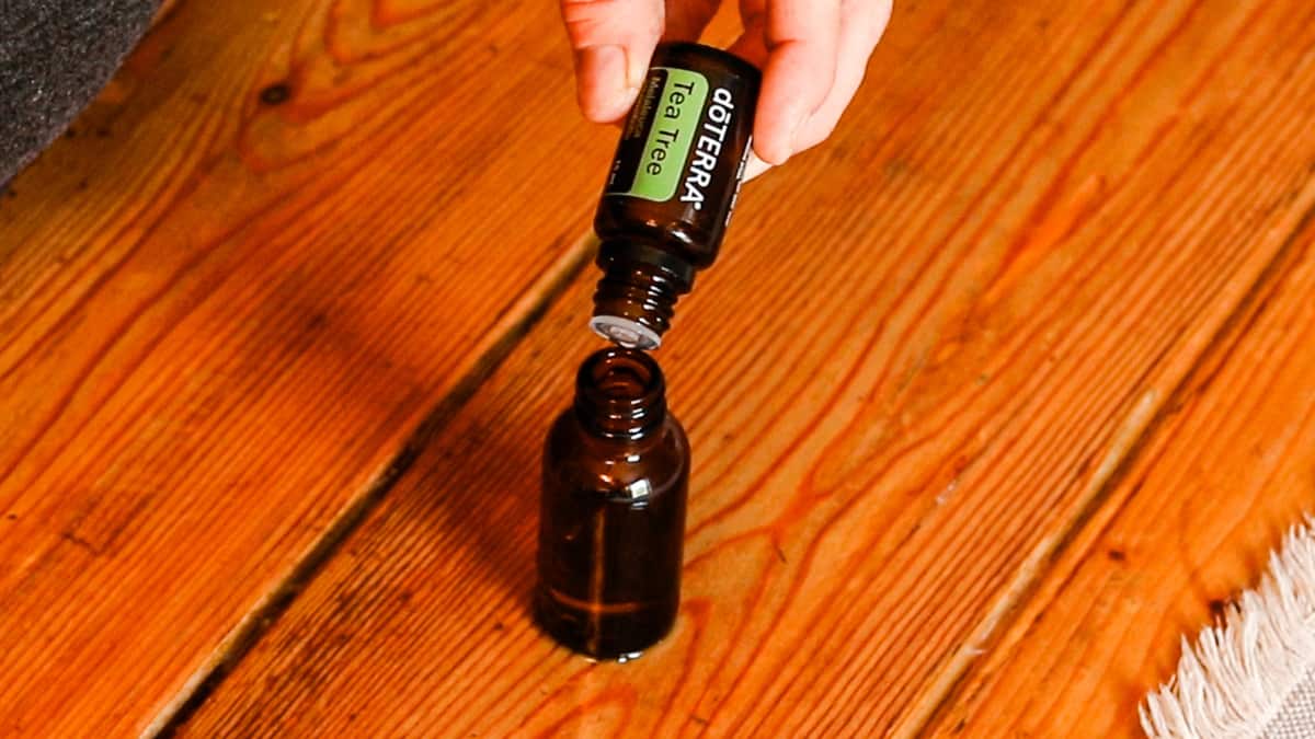 A hand is dropping doTERRA tea tree oil into a brown glass dropper bottle. The bottle sits on a wooden table with an ivory table runner.