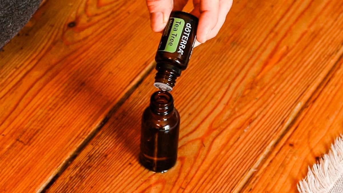 A hand is dropping doTERRA tea tree oil into a brown glass dropper bottle. The bottle sits on a wooden table with an ivory table runner.