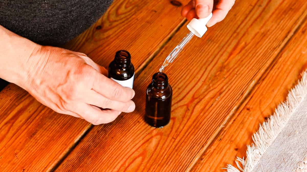 Two hands are dropping hyaluronic acid into a brown, glass dropper bottle. The bottle sits on a wooden table with an ivory table runner.