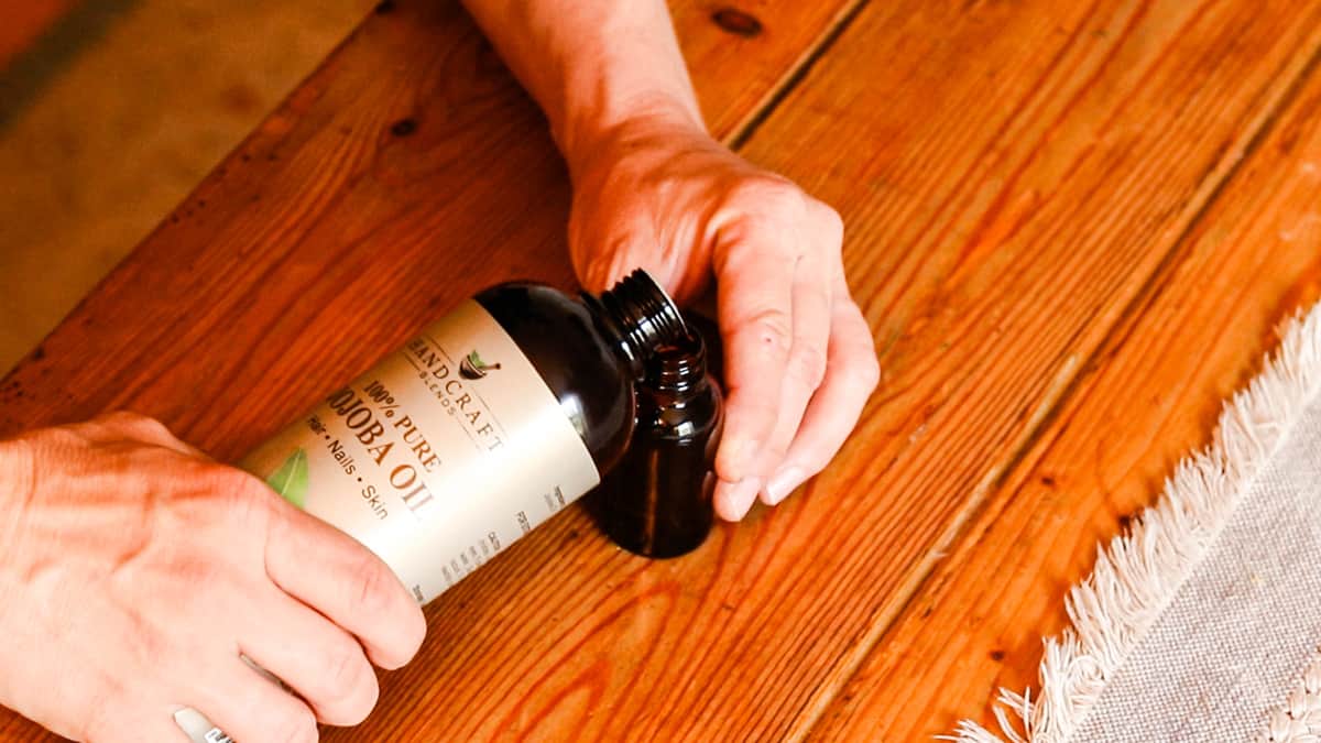 Two hands are pouring jojoba oil into a small, dark glass dropper bottle. There is a wooden table and an ivory table runner.