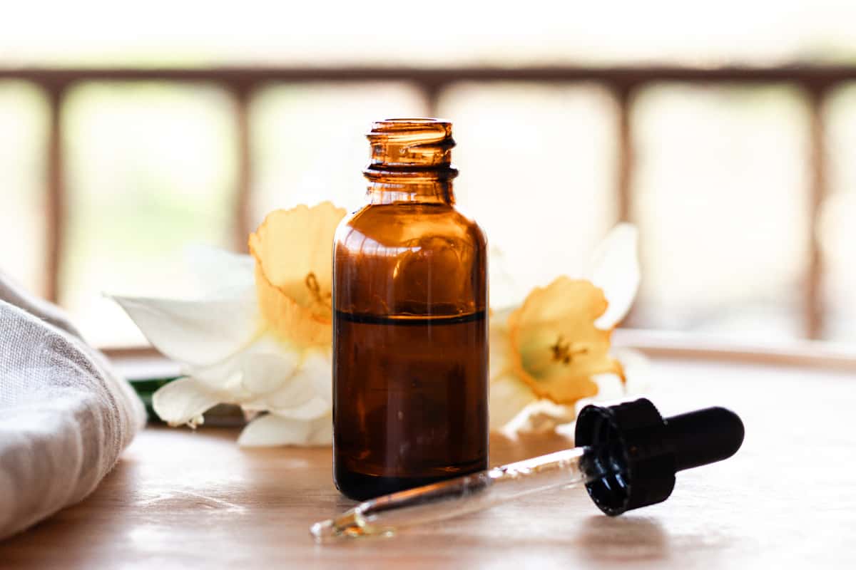 A brown glass bottle filled with homemade dry scalp serum. The dropper lid lies on the table in front and there is a daffodil in the background for decoration. 