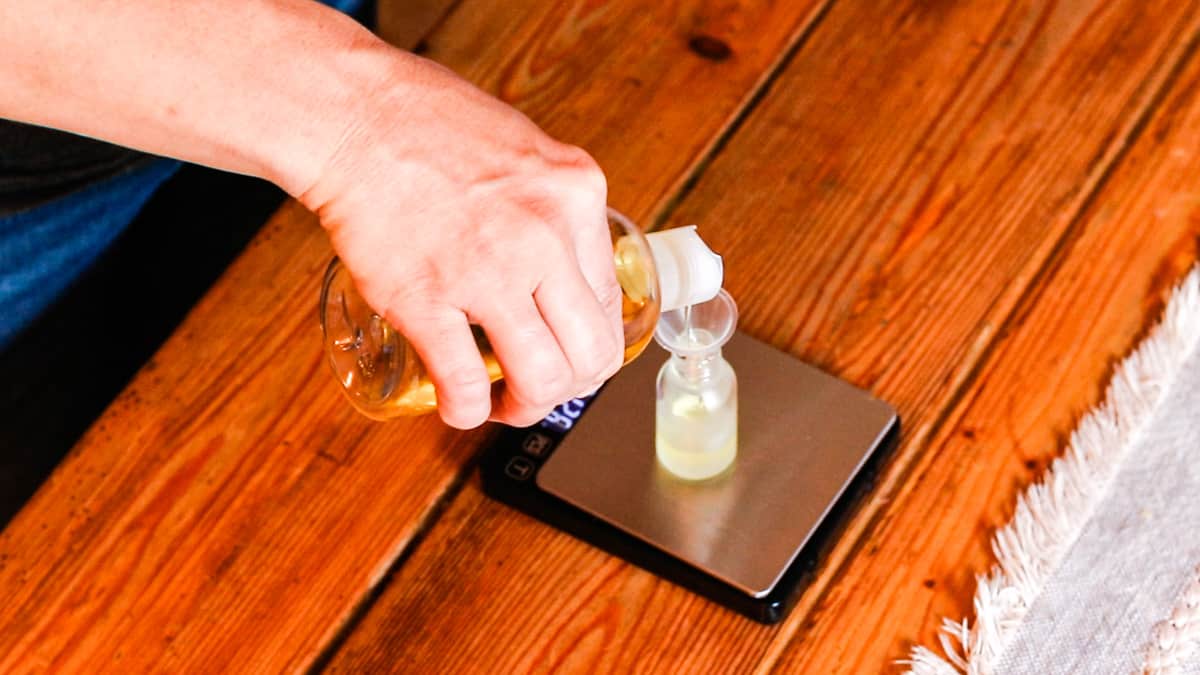 A hand pouring a carrier oil into a small glass bottle using a small funnel. The glass bottle is on a digital scale which is resting on a wooden work table. The mixture is part of a DIY face serum for youthful skin. 