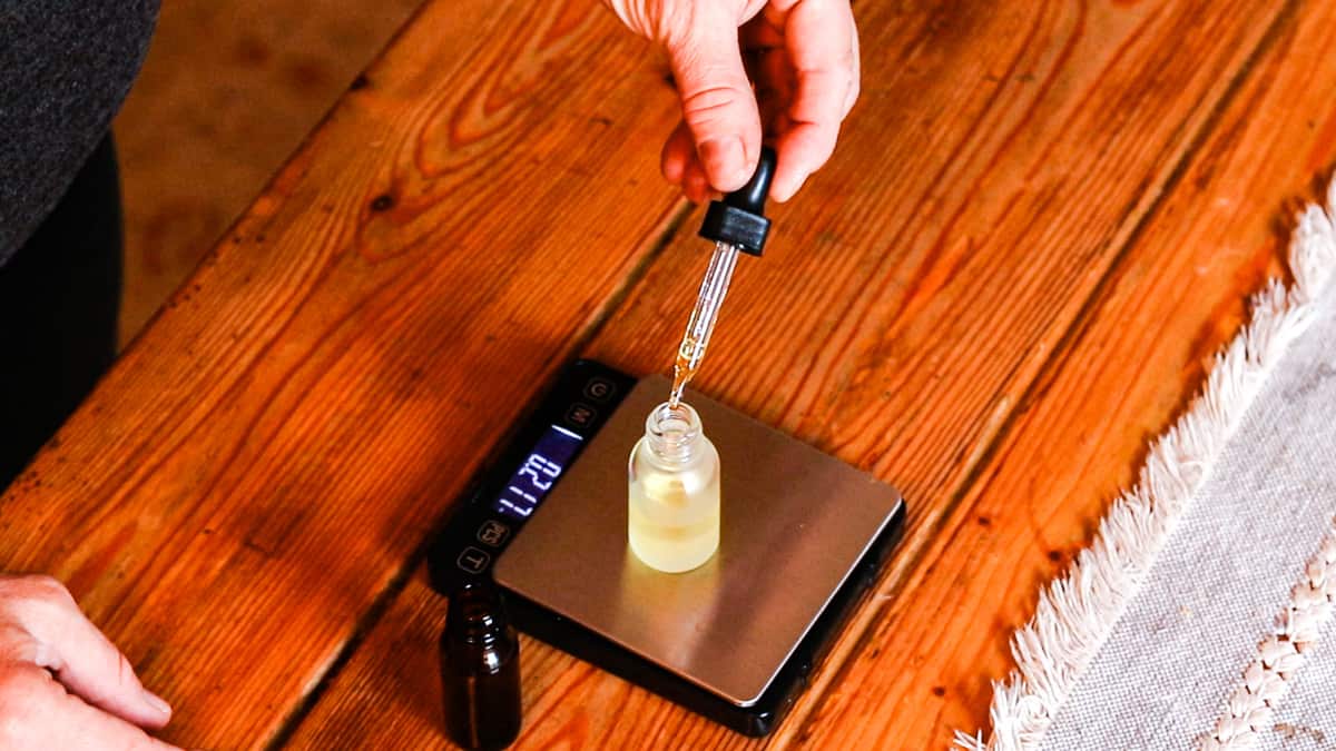 A glass bottle on a scale with Vitamin E being dropped into it. Digital metal scale rests on a wooden table. Bottle holds a DIY facial serum for anti-aging.