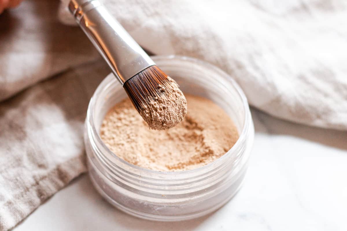 A makeup brush dipped in mineral makeup with a small jar of homemade mineral makeup in the background.